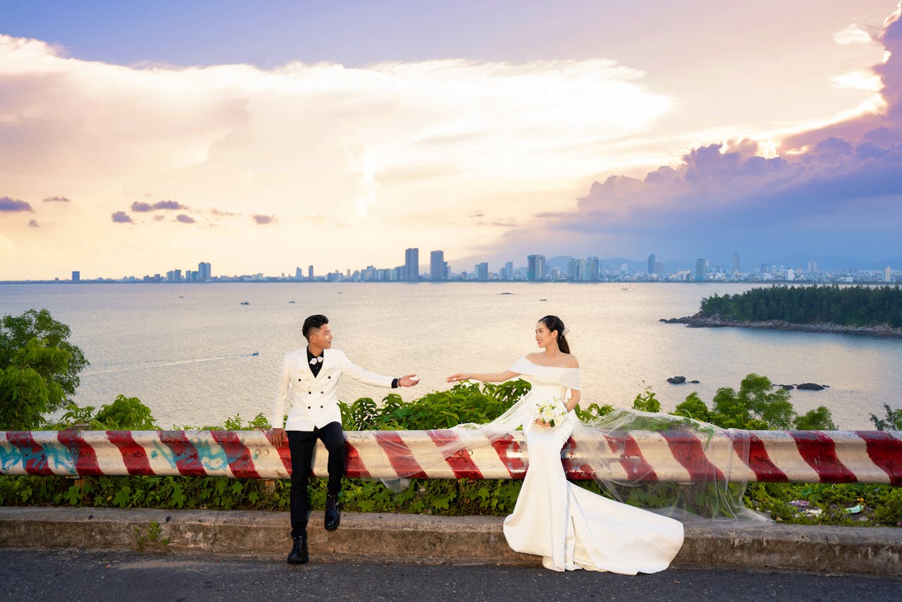 A couple poses for wedding photos by the sea at sunset with a city skyline in the distance.